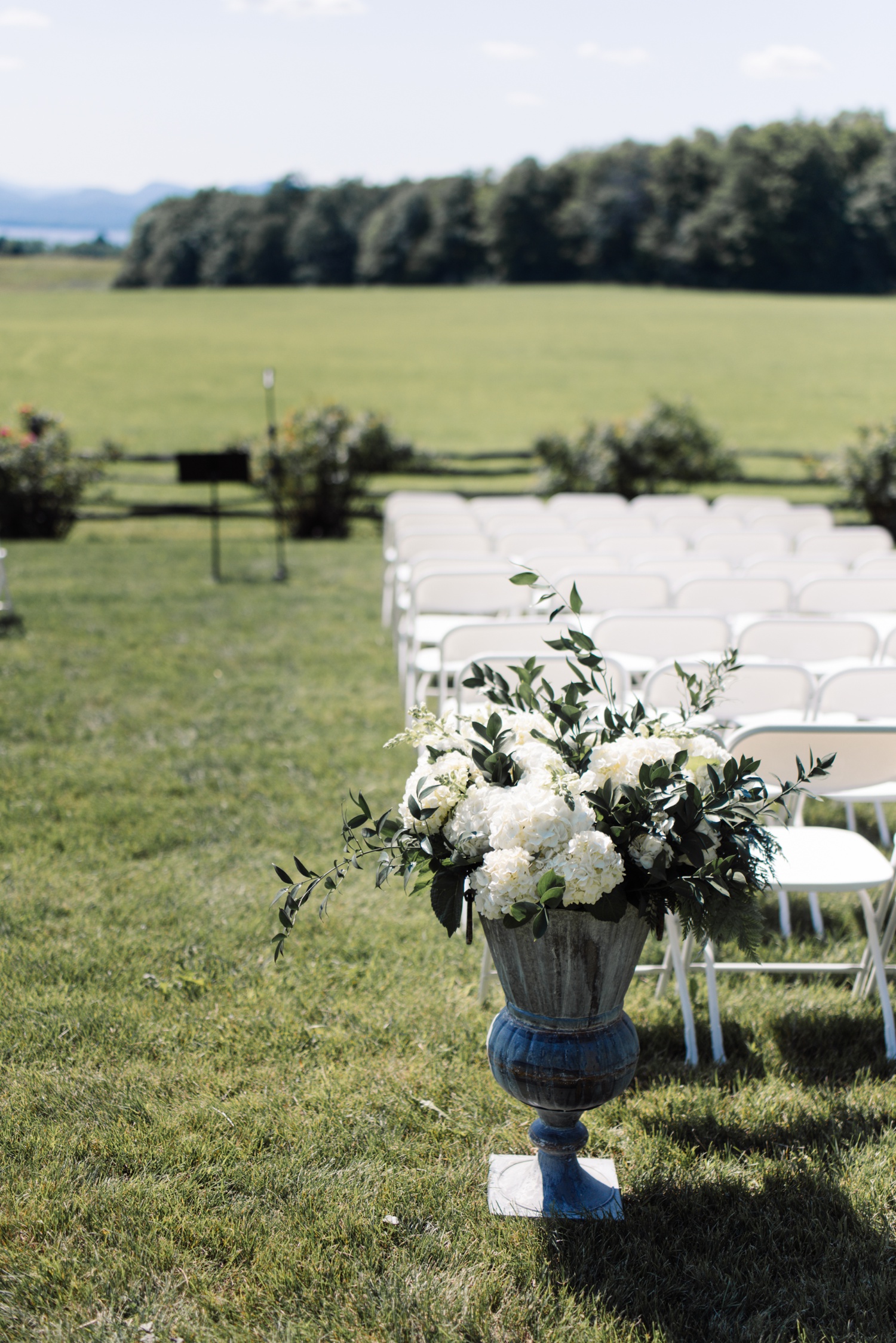 White folding chairs and a large vase filled with white hydrangeas and greenery for a summer outdoor wedding ceremony