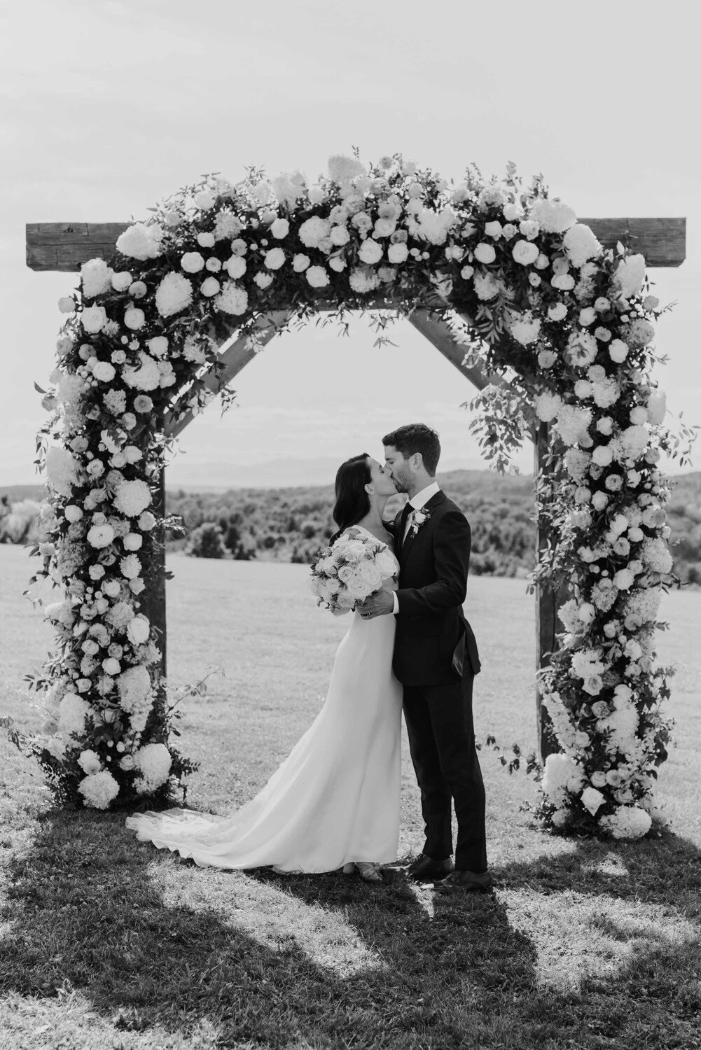 Bride and groom kiss for first time greyscale, outdoors summer inspired in real wedding stevie gown by alexandra grecco