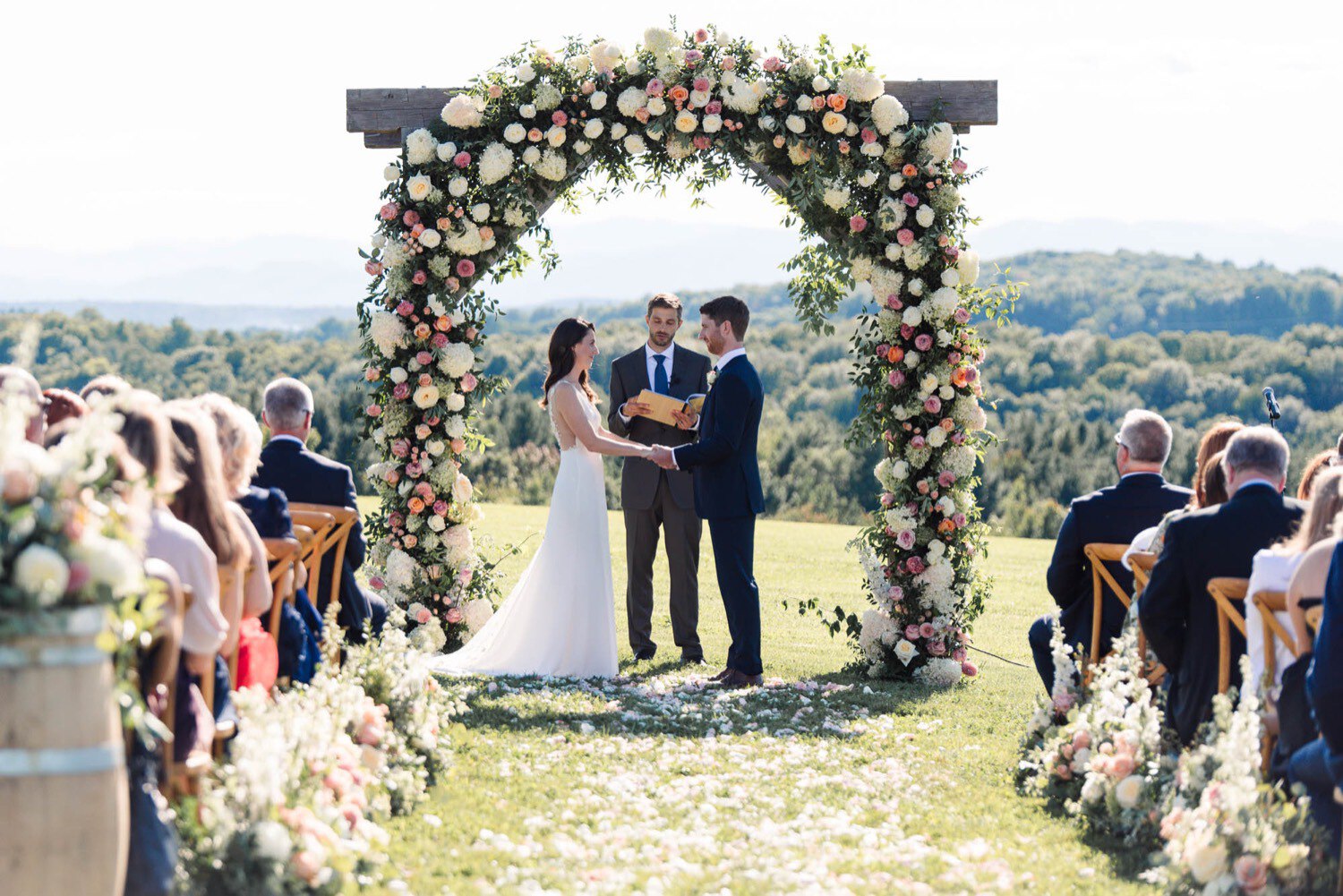 Officiant weds couple in outdoor summer wedding ceremony with lush floral arch by clayton floral