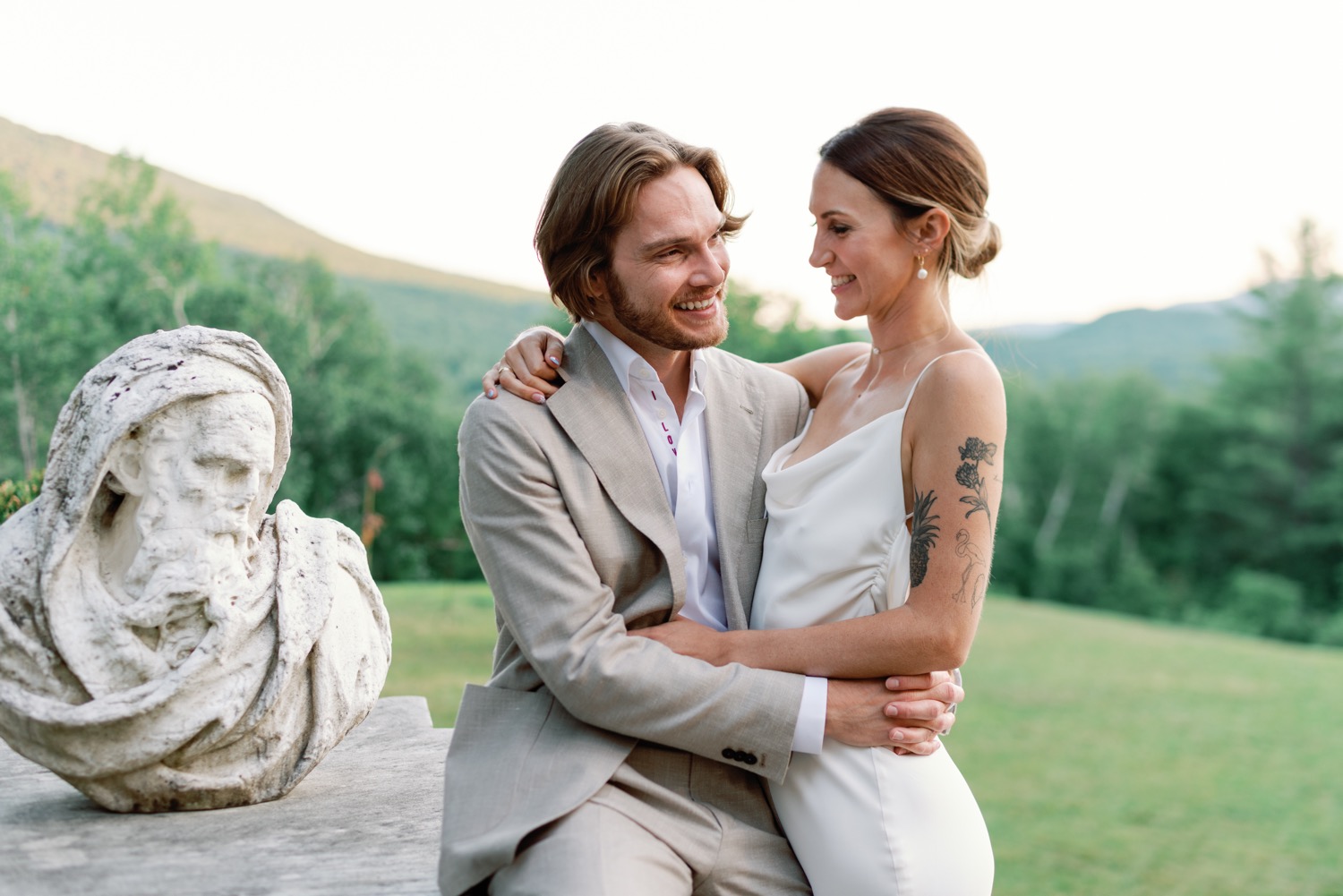 Newlyweds embrace sitting next to bust statue at wilburton Inn manchester vt with silky alexandra grecco gown and groom in pini parma suit unique wedding idea