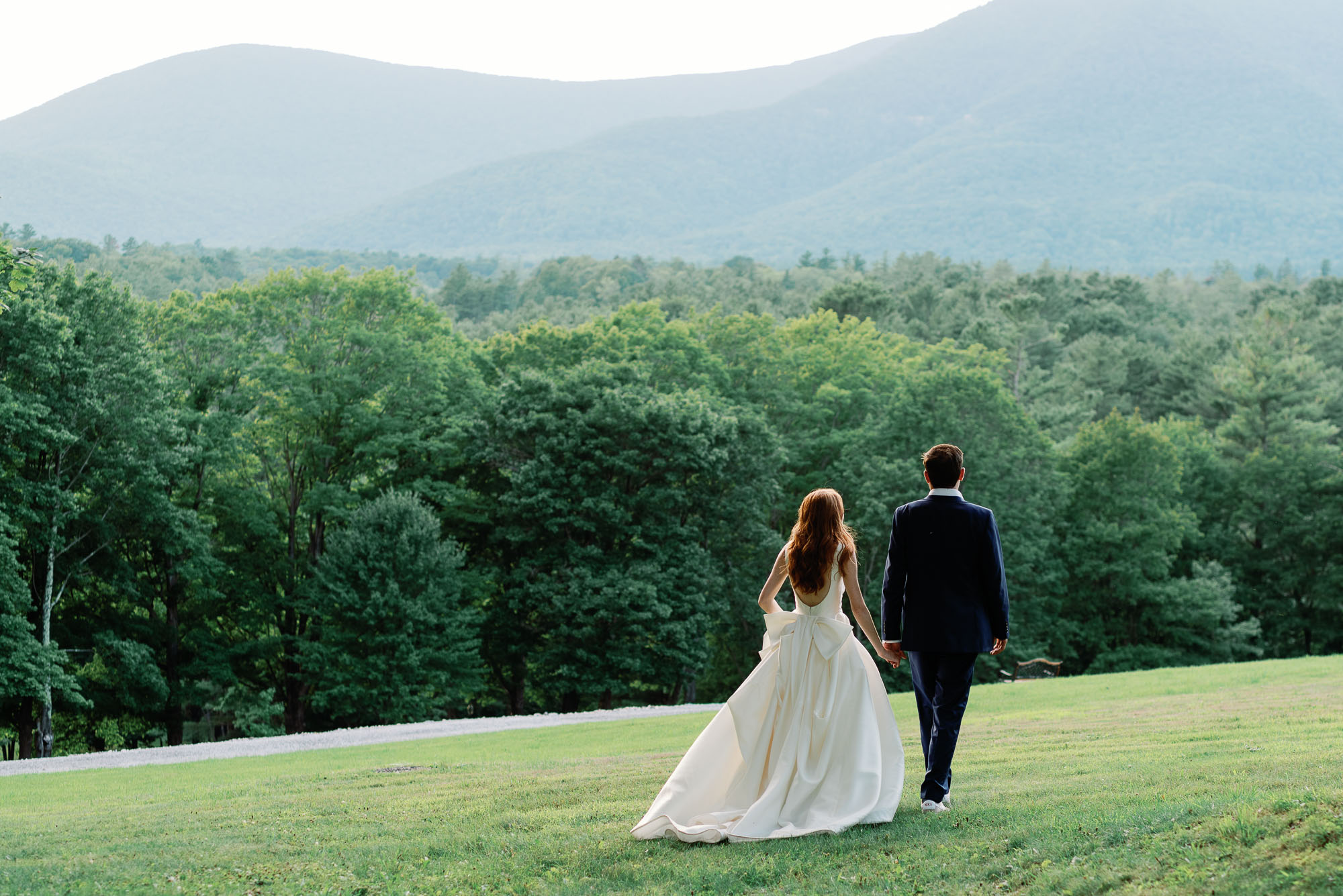 wedding couple walks across green grass with mountains in the background at the wilburton inn in vermont at elegant new england wedding