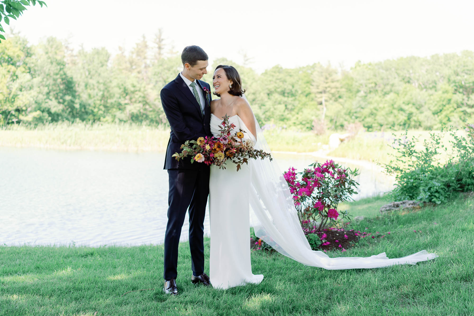 sarah seven bride smiles at groom with pink bouquet next to pond in vermont