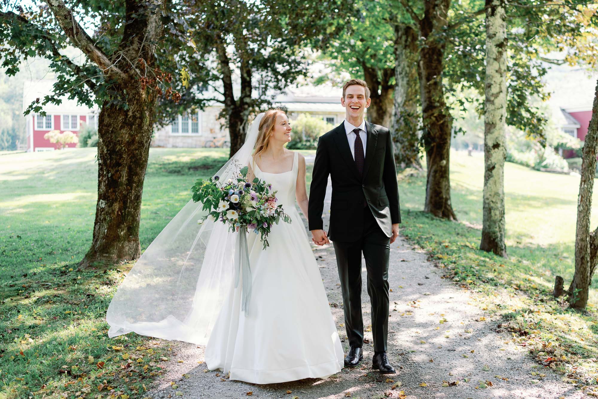 bride and groom at riverside farm wedding in vermont walk down forested pathway smiling and laughing in new england town